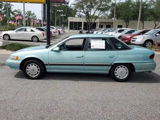 Passenger-side profile of the light teal 1994 Mercury Sable GS four-door sedan on a dealer lot