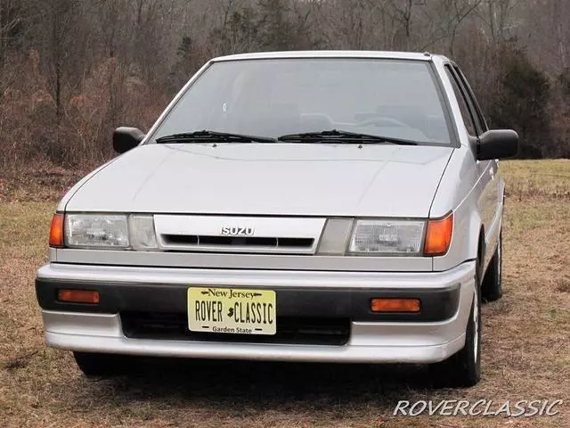 Front view of a silver 1989 Isuzu I-Mark parked on dry grass in front of bare trees, with a New Jersey dealer plate