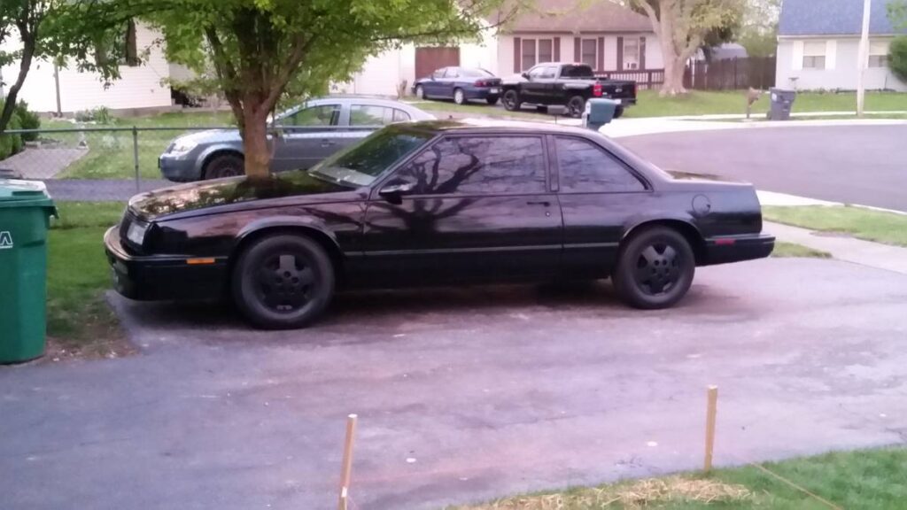Black 1987 Buick LeSabre T-Type coupe with black wheels parked in a driveway, front three-quarter view