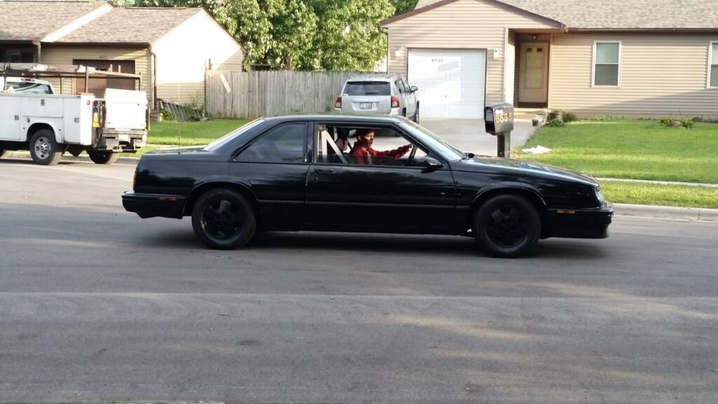 Driver-side profile of the black 1987 Buick LeSabre T-Type coupe being driven on a residential street