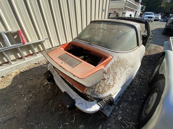 Stripped white 1979 Porsche 911 Targa shell beside a metal building, rear view with the engine lid open and no engine fitted