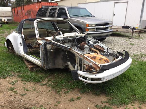 Front three-quarter view of the stripped white 1979 Porsche 911 Targa shell on grass, with heavy rust in the front trunk area and the interior gutted